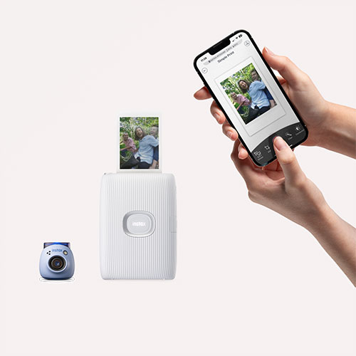 Mani tengono un telefono con foto, una stampante bianca stampa la foto, una fotocamera blu.