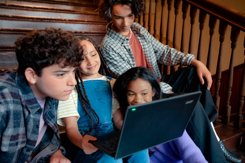 Quattro ragazzi guardano un laptop su una scala di legno. Indossano vestiti casual e sembrano impegnati con lo schermo.