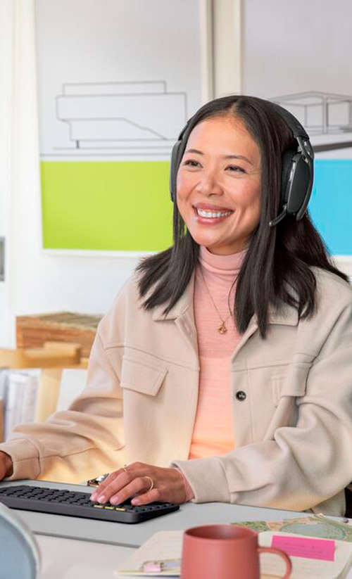 Donna sorridente con cuffie lavora al computer. Tazza rosa sul tavolo, sfondo con disegno architettonico e aree colorate.