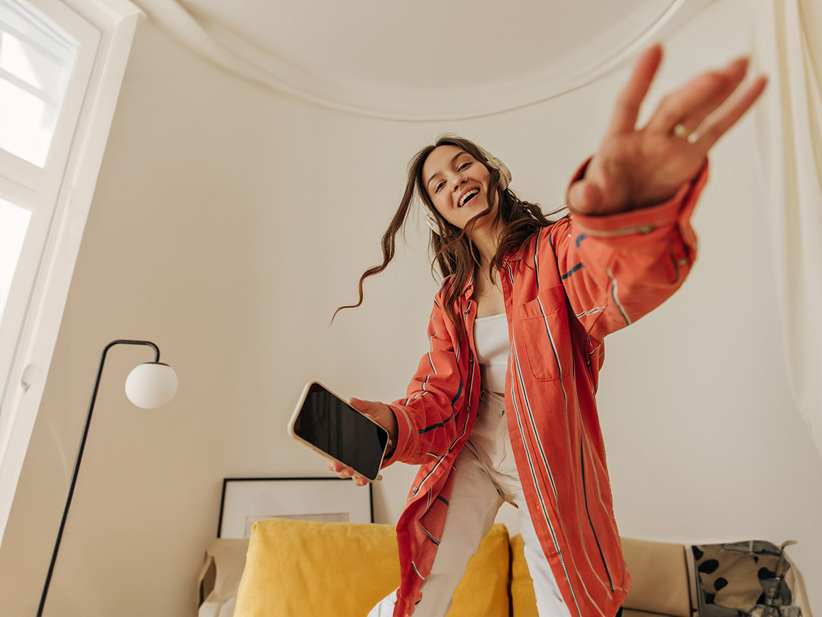 Donna sorridente con cuffie, camicia a righe arancione, pantaloni bianchi tiene un telefono in una stanza chiara.