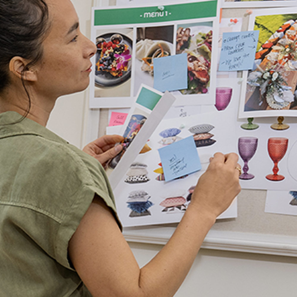 Donna in camicia verde lavora a un pannello con immagini di cibo, cuscini e bicchieri, usando appunti adesivi.