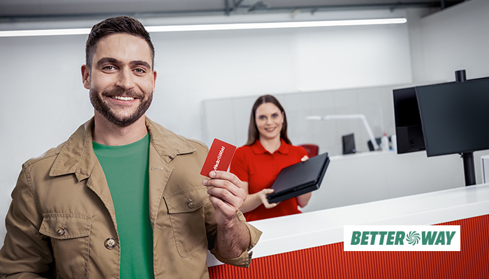 Uomo sorridente con carta rossa, donna con laptop rosso dietro un bancone bianco. Logo "Better Way".