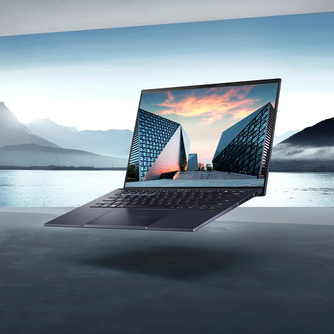 Un laptop nero aperto mostra edifici moderni su uno sfondo di lago e montagne. Il laptop è su una superficie grigia.