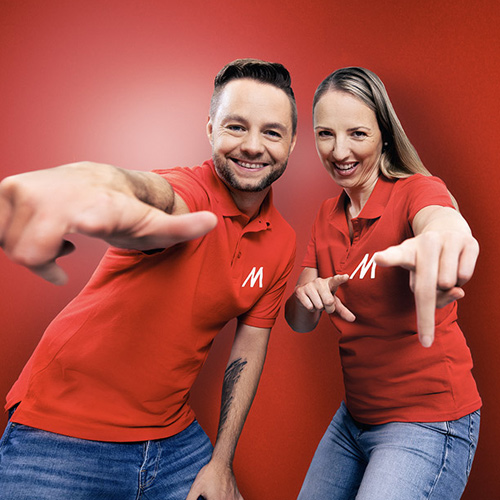 Due persone sorridono, indossando polo rosse con "M", puntando verso la fotocamera su sfondo rosso. Indossano jeans.