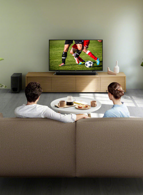 Due persone sedute su un divano guardano una partita di calcio in TV. Un tavolino con tazze e cibo � davanti a loro.