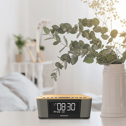 Sveglia digitale Kenwood su un tavolo, che mostra l'ora 08:30. Sullo sfondo, un vaso bianco con rami e un letto.