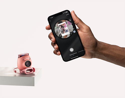 Una mano tiene un telefono che fotografa una fotocamera rosa su un cubo bianco. Lo sfondo � bianco.