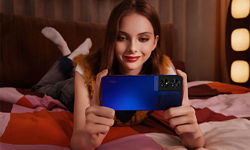 Donna distesa su un letto a motivi rossi e bianchi, tiene un telefono blu orizzontalmente. Sfondo: lampada con luci.