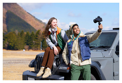 Due persone accanto a un'auto grigia. Una persona tiene in mano una videocamera. Alberi e montagne sullo sfondo.