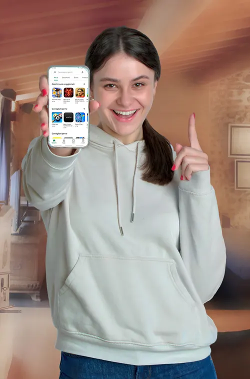 Donna sorridente mostra uno smartphone con app, indossa felpa grigia e jeans blu. Dietro, parete marrone con quadri.