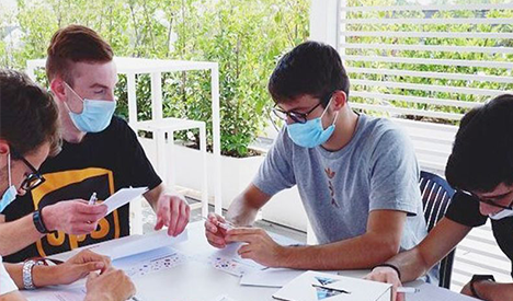 Persone sedute a un tavolo all'aperto con maschere, che lavorano su documenti. Sul tavolo ci sono carta e penne.