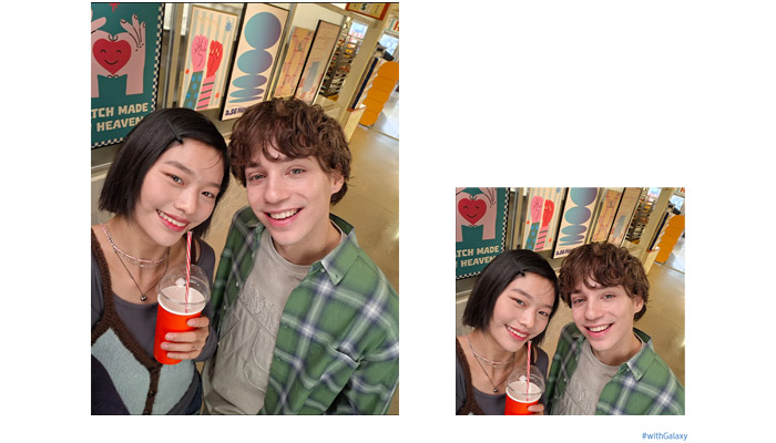 Due persone sorridono, una tiene una bevanda. Dietro, cartelli con cuori rossi e scritte. Uno indossa una camicia a quadri verde.