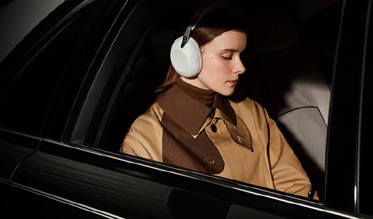 Una donna seduta sul sedile posteriore di un'auto scura indossa grandi cuffie over-ear bianche, con gli occhi socchiusi e un cappotto marrone e beige.
