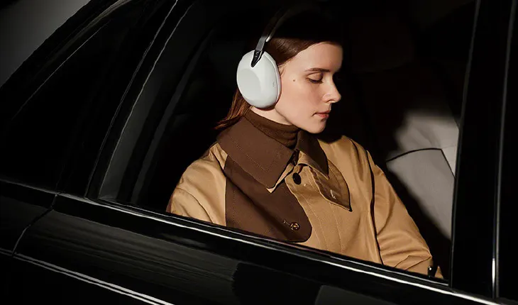 Una donna seduta sul sedile posteriore di un'auto scura indossa grandi cuffie over-ear bianche, con gli occhi socchiusi e un cappotto marrone e beige.