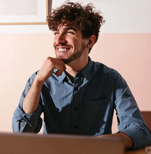 Uomo sorridente con camicia blu e auricolare. Quadro appeso al muro color pesca in secondo piano. Parte di un laptop in primo piano.