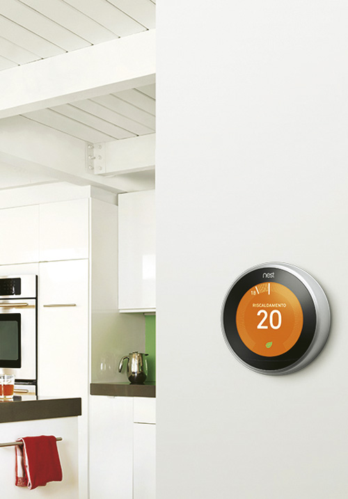 Termostato Nest su muro bianco, indica 20 gradi. In secondo piano, cucina bianca con elettrodomestici e accessori.