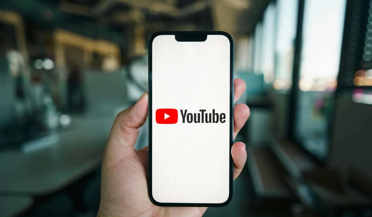 Una mano tiene un telefono che mostra il logo di YouTube su uno sfondo luminoso e sfocato.