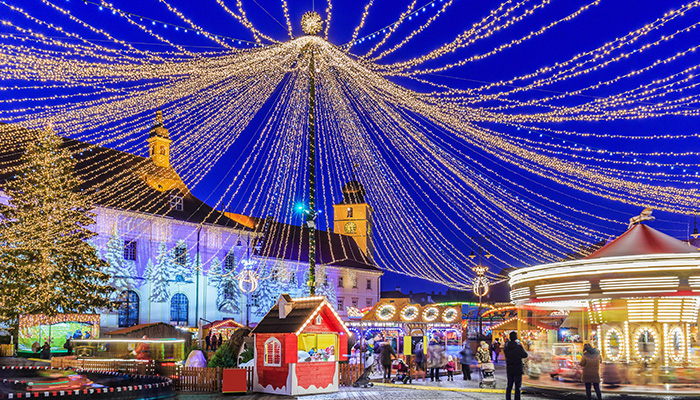 Mercato di Natale notturno con giostra, bancarelle rosse e persone. Il cielo blu è decorato con luci dorate.