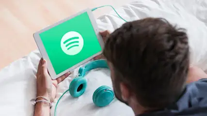 Uomo a letto con cuffie turchesi. Un tablet mostra il logo Spotify. Lenzuola bianche e pavimento chiaro.