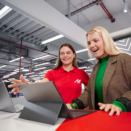 Due donne guardano dei laptop. Una indossa una polo rossa con una "M", l'altra una giacca a quadri marrone e una maglia verde.