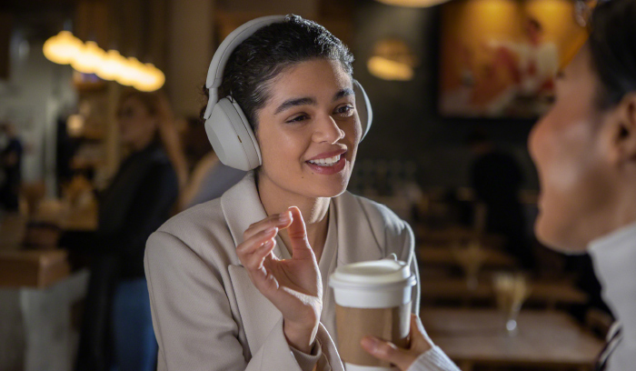 Donna con cuffie bianche, giacca beige, tiene tazza di caff�. Sullo sfondo, luci soffuse e altre persone in un bar.