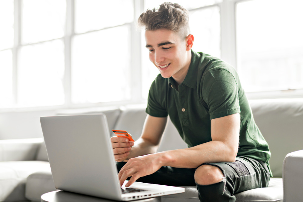 Un uomo usa un portatile, tenendo una carta arancione. Indossa una polo verde e pantaloni neri strappati. Sullo sfondo, un divano grigio.