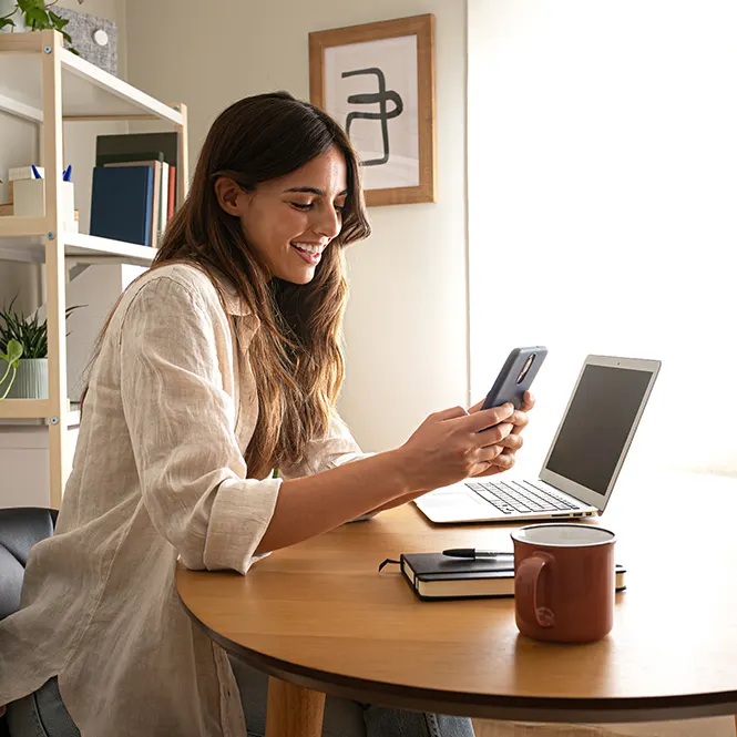 Donna sorridente usa uno smartphone vicino a un laptop su un tavolo di legno con tazza e blocco note. Una libreria � sullo sfondo.