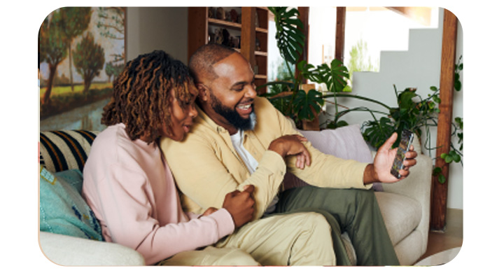 Due persone sedute su un divano usano un telefono. Piante e una scala sono sullo sfondo.