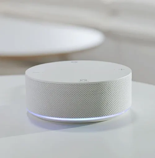 Dispositivo circolare bianco con griglia laterale e luce blu inferiore su superficie chiara. Tavolo rotondo sfocato sullo sfondo.