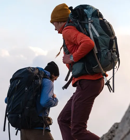 Due escursionisti con zaini camminano in salita. Uno indossa un cappello giallo e giacca arancione, l'altro un cappello nero e giacca blu.