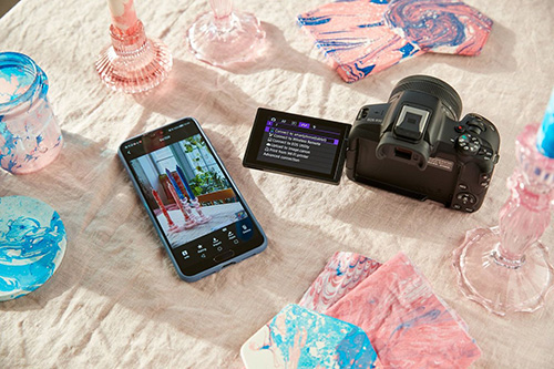 Su un tessuto chiaro sono disposti una fotocamera nera, un telefono, un tablet e oggetti decorativi blu e rosa.