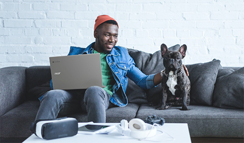 Uomo seduto su un divano grigio con un laptop. Un cane nero � seduto accanto a lui. Un tavolo bianco ha cuffie e un controller.