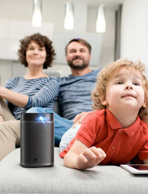 Famiglia su divano grigio con bambino in primo piano. Un proiettore nero è visibile accanto al bambino. Lampade a sospensione sullo sfondo.