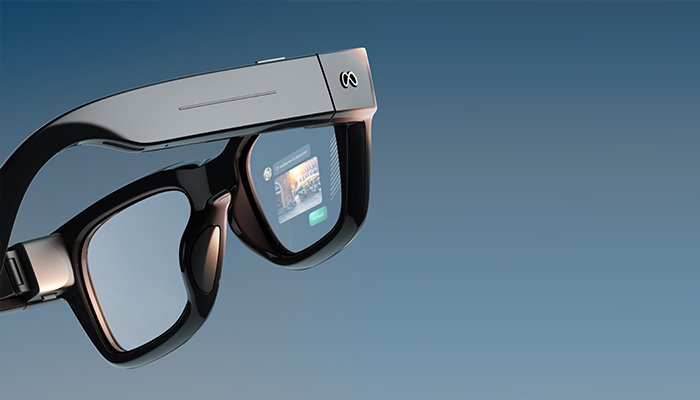Primo piano di occhiali smart neri con montatura squadrata e display integrato nella lente destra, mostrati su uno sfondo sfumato blu-grigio.