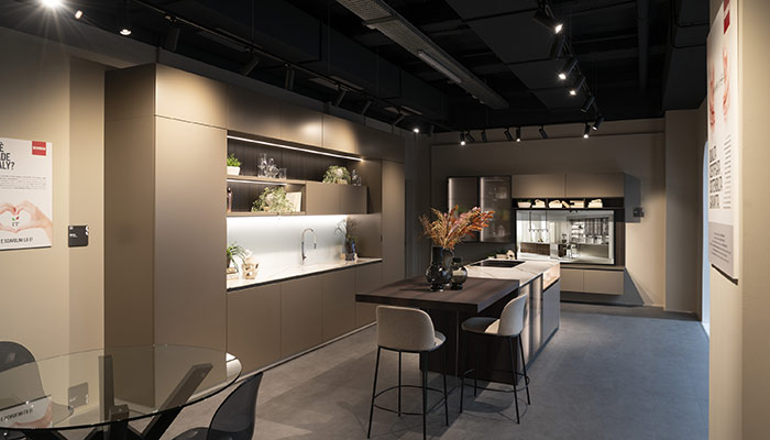 Interno di una cucina moderna con isola centrale, sgabelli, ripiani e scaffali illuminati.