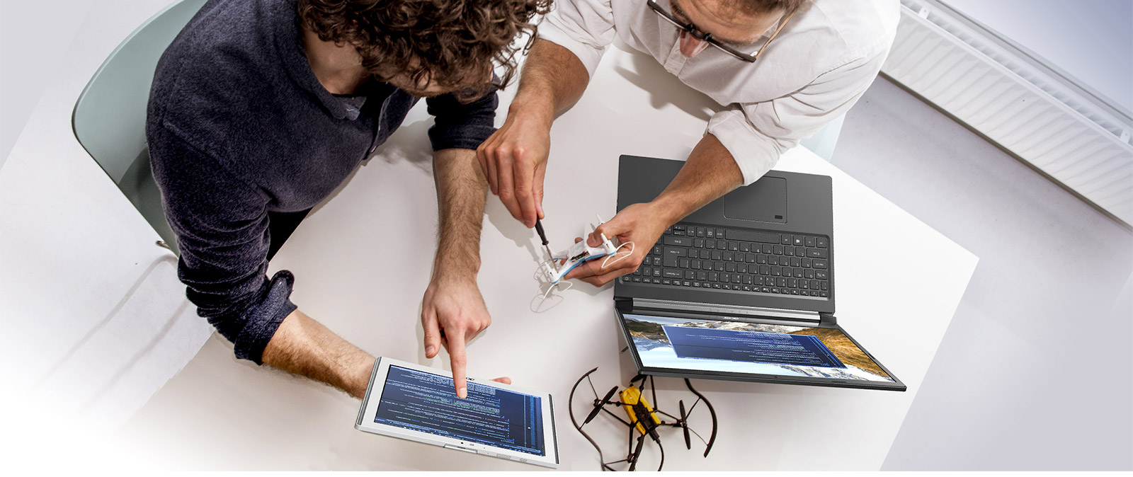 Due persone lavorano su un drone giallo su un tavolo bianco con un laptop e un tablet che mostrano codice.