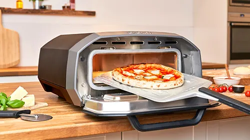 Forno per pizza con pizza su pala, formaggio, basilico e tagliere in legno su ripiano in cucina.