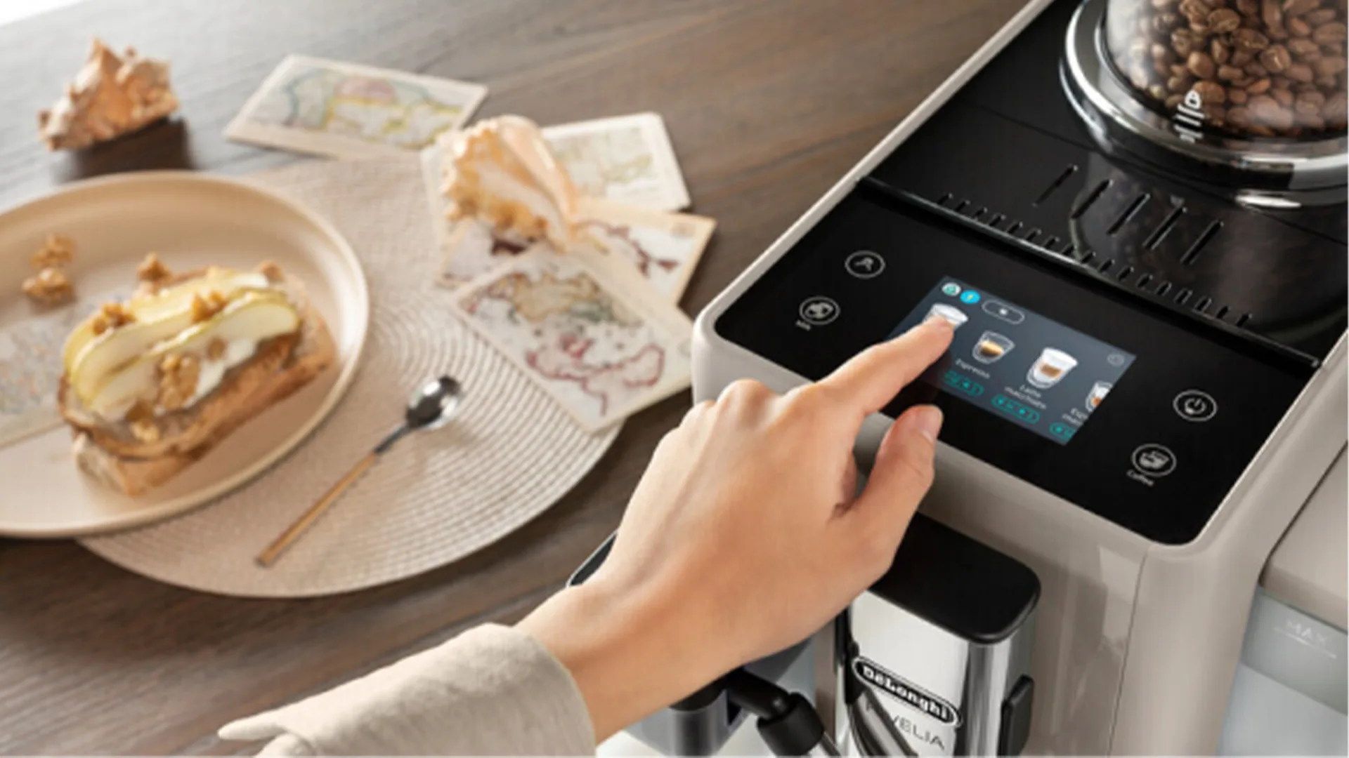 Una mano tocca lo schermo di una macchina da caff�. Sul tavolo, un piatto con pane tostato, una conchiglia e delle cartoline.