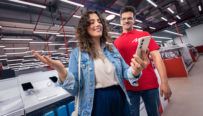 Donna in negozio con giacca di jeans accanto a uomo in maglietta rossa che tiene un telefono. Espositori in secondo piano.