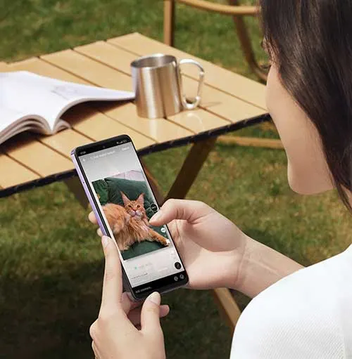 Donna con telefono che mostra foto di gatto su tavolo in legno con libro e tazza in metallo, su prato.