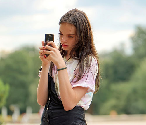Ragazza tiene un telefono cellulare. Indossa una maglietta bianca e una salopette nera. Sullo sfondo alberi e cielo.