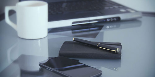 Tavolo con laptop, tazza bianca, blocco note nero, penna e cellulare. Gli oggetti sono disposti in modo ordinato.