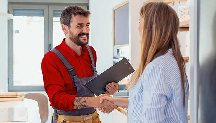 Un idraulico in tuta rossa stringe la mano a una donna in camicia a righe in una stanza con finestra.