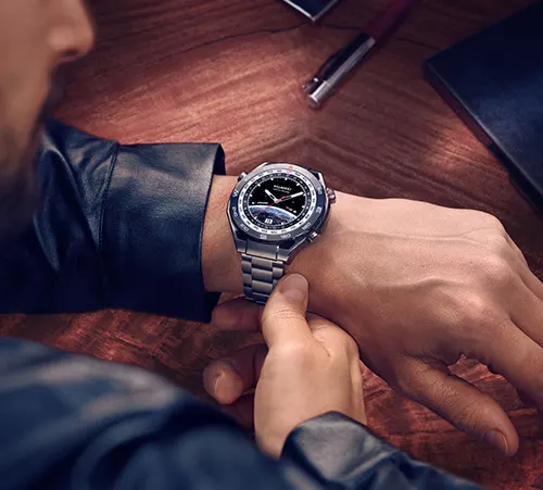 Un uomo indossa un orologio da polso in metallo. Sul tavolo di legno ci sono una penna, un cellulare e un tablet nero.