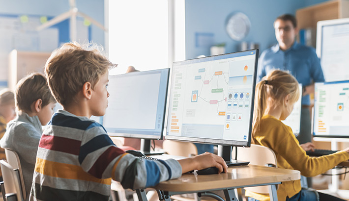 Ragazzi in aula informatica, concentrati sui computer. Un insegnante in piedi sullo sfondo. Ambiente scolastico luminoso e moderno.