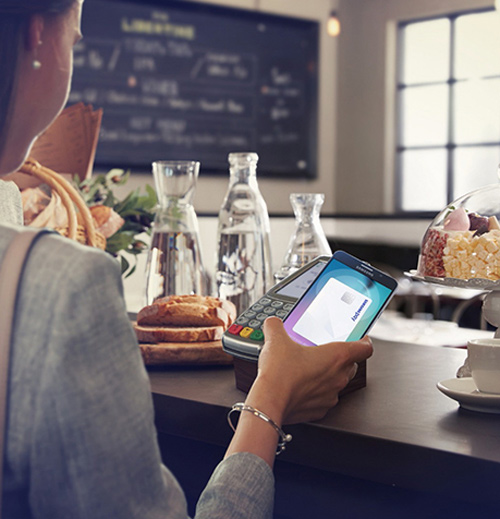 Donna usa un terminale di pagamento in un bar con dolci, pane e caraffe sullo sfondo.