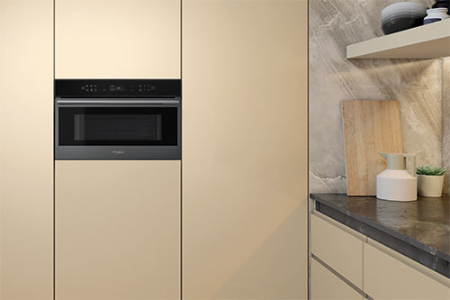 Cucina con forno integrato in mobili beige. Un ripiano con ciotole, tagliere, vaso bianco e pianta decorativa.