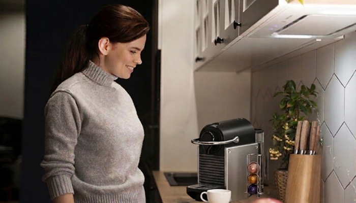Una donna sorridente in maglione grigio guarda una macchina del caffè a capsule sul bancone della cucina.