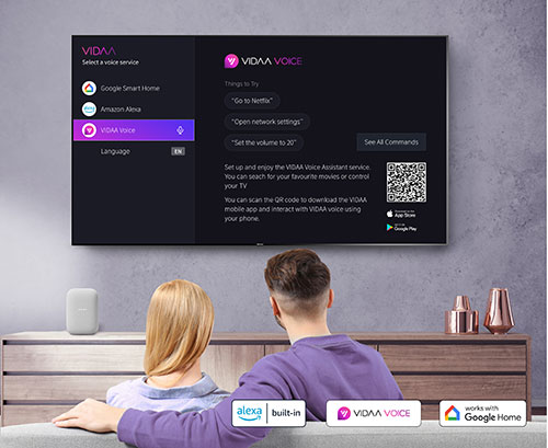 Coppia davanti a TV che mostra il menu vocale VIDA, con logo Alexa, Google Home e altoparlante su mobiletto.