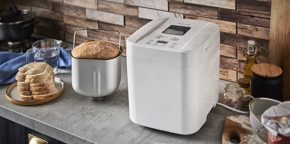 Cucina con macchina per il pane bianca, pane in cassetta, brocca d'acqua e utensili su un piano di lavoro grigio.